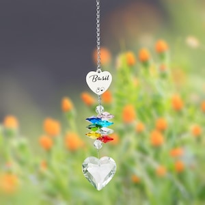 Puede incluir: Un atrapasoles de cristal en forma de coraz&oacute;n, con el nombre "Basil" inscrito en un peque&ntilde;o dije en forma de coraz&oacute;n. Cuenta con una cadena, cristales de colores y un cristal grande en forma de coraz&oacute;n en la parte inferior. El fondo son flores naranjas borrosas.