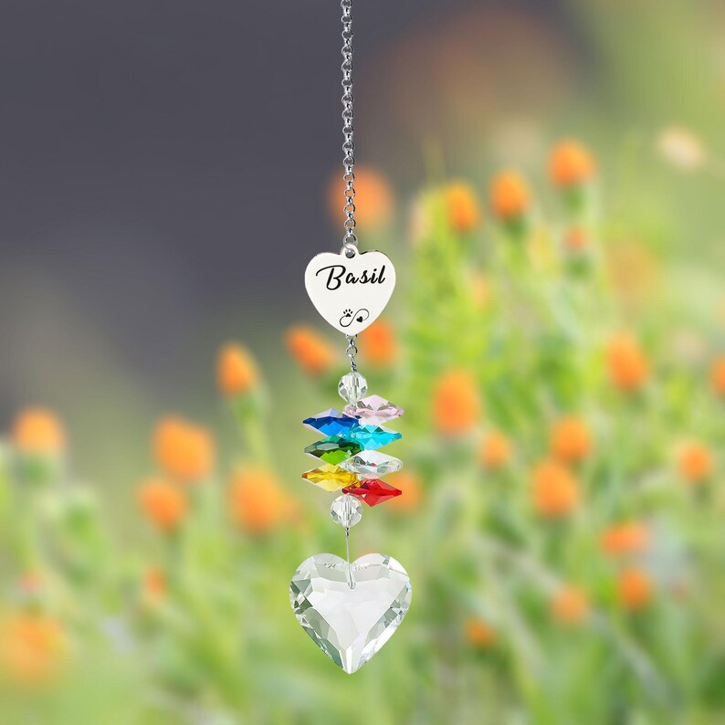 Puede incluir: Un atrapasoles de cristal en forma de coraz&oacute;n, con el nombre "Basil" inscrito en un peque&ntilde;o dije en forma de coraz&oacute;n. Cuenta con una cadena, cristales de colores y un cristal grande en forma de coraz&oacute;n en la parte inferior. El fondo son flores naranjas borrosas.