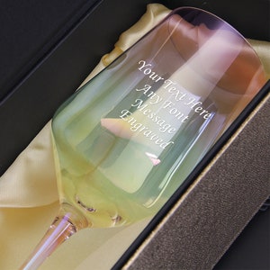 May include: A clear glass wine glass with a rainbow iridescent finish. The glass is engraved with the text "Your Text Here Any Font Message Engraved". The glass is in a black box with a gold lining.