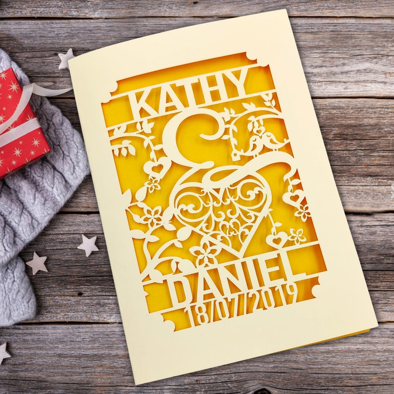 May include: A yellow and cream card with a cut-out design featuring a heart, flowers, birds, and the names "Kathy" and "Daniel" with the date "18/07/2019".