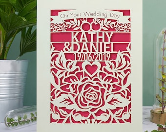 May include: A white card with a pink floral design and the text "On Your Wedding Day" with the names "Kathy & Daniel" and the date "19/06/2019".