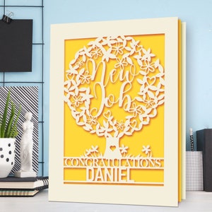 May include: A white card with a yellow background and a cut-out design of a tree with the words "New Job" inside the tree. The card also has the words "Congratulations Daniel" at the bottom.