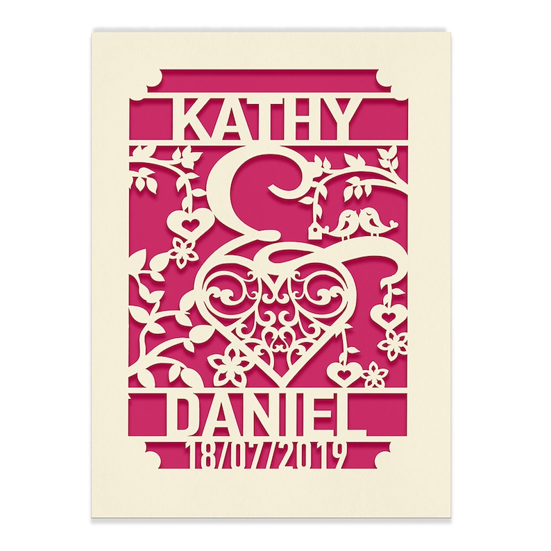 May include: A white paper cut-out design with a pink background. The design features a heart with a decorative flourish, two birds, and the names "Kathy & Daniel" with the date "18/07/2019".