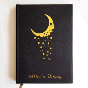May include: A black hardcover journal featuring a gold foil design of a crescent moon with a trail of falling stars. The text Alice's Diary is printed in gold script at the bottom. The cover has a textured finish and a gold ribbon bookmark is visible.