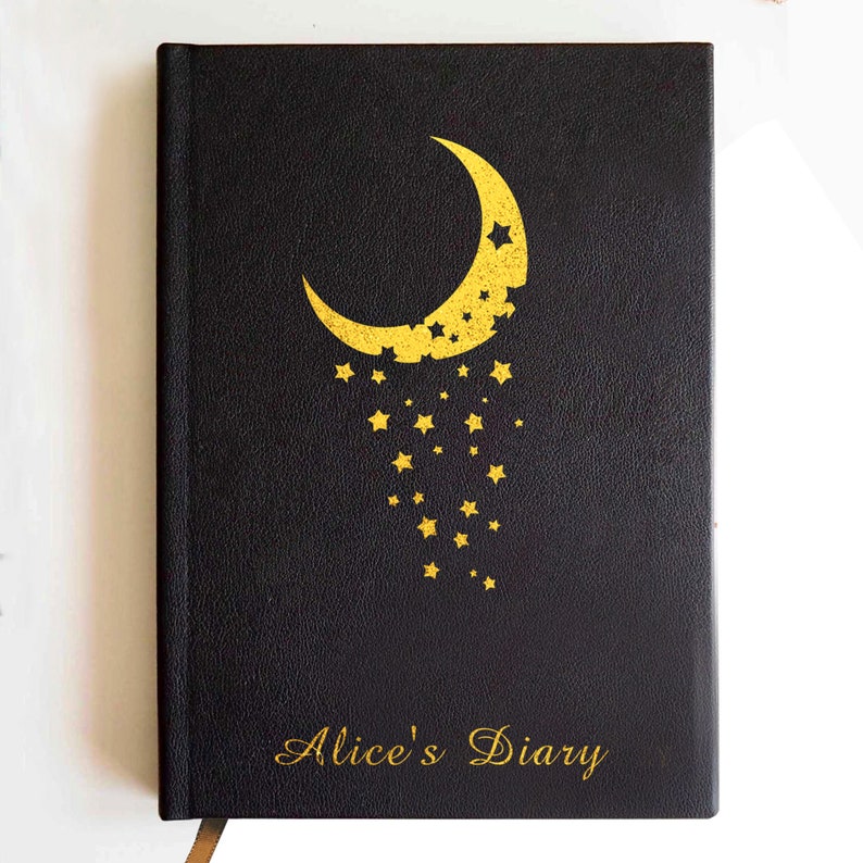 May include: A black hardcover journal featuring a gold foil design of a crescent moon with a trail of falling stars. The text Alice's Diary is printed in gold script at the bottom. The cover has a textured finish and a gold ribbon bookmark is visible.