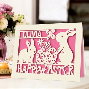 May include: A white card with a pink background featuring a cut-out design of two bunnies, a basket of Easter eggs, and flowers. The card says "Olivia Happy Easter".