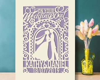 May include: A white paper wedding invitation with a purple background. The invitation features a cut-out design of a couple standing under an archway with flowers and the text "On Your Wedding Day". The names "Kathy & Daniel" and the date "18/07/2019" are also included.