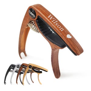 May include: A wooden capo with the name "Wilson" and a heart symbol printed on it. The capo has a black rubber grip and a silver spring. Several other capos in black, silver, brown, and light brown are displayed below.
