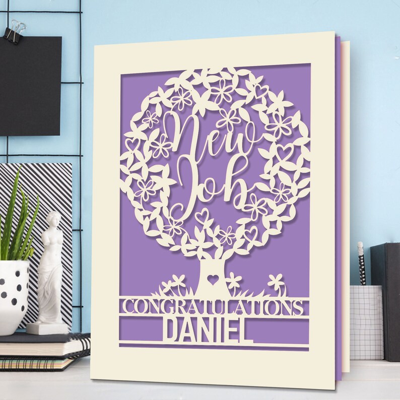 May include: A white card with a purple background and a white paper cut-out design of a tree with the words "New Job" inside the tree. The card also has the words "Congratulations Daniel" at the bottom.