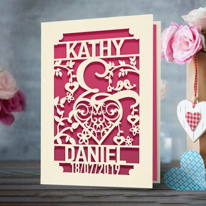 May include: A cream-colored card with a pink cutout design. The design features a heart with two birds perched on a branch, surrounded by flowers and leaves. The names "Kathy & Daniel" and the date "18/07/2019" are cut out below the heart.