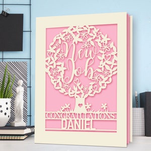 May include: A cream-colored card with a pink background features a white paper cut-out design of a tree with leaves and flowers. The tree has the words "New Job" inside the leaves. The card also has the words "Congratulations Daniel" at the bottom.