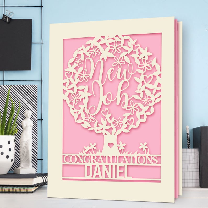 May include: A cream-colored card with a pink background features a white paper cut-out design of a tree with leaves and flowers. The tree has the words "New Job" inside the leaves. The card also has the words "Congratulations Daniel" at the bottom.