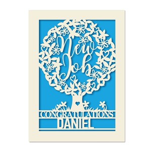 May include: A white card with a blue background. The card features a white paper cut-out design of a tree with the words "New Job" inside the tree. The text "Congratulations Daniel" is below the tree.