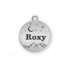 Puede incluir: Una placa redonda y plateada para mascotas con el nombre "Roxy" en letras negras. El dise&ntilde;o incluye una cordillera, una luna creciente y estrellas. Un peque&ntilde;o lazo en la parte superior.