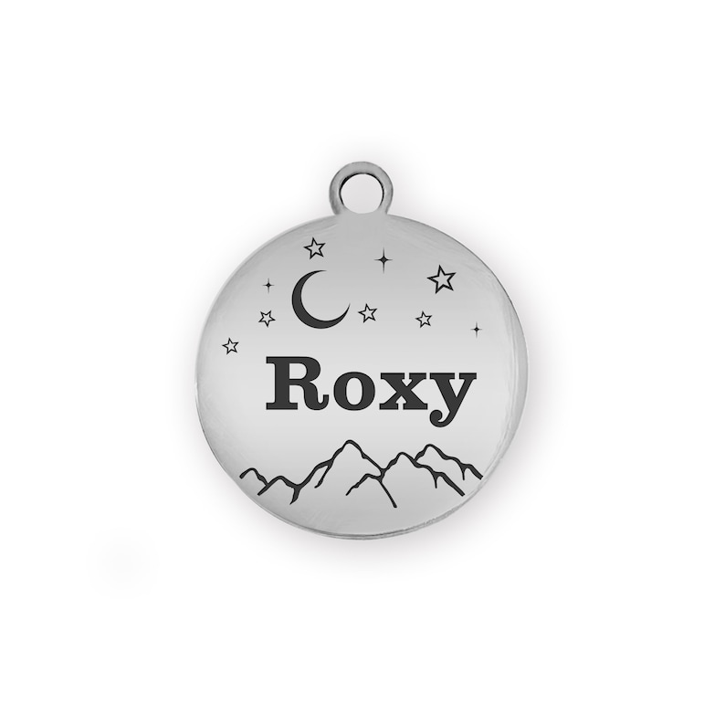 Puede incluir: Una placa redonda y plateada para mascotas con el nombre "Roxy" en letras negras. El dise&ntilde;o incluye una cordillera, una luna creciente y estrellas. Un peque&ntilde;o lazo en la parte superior.