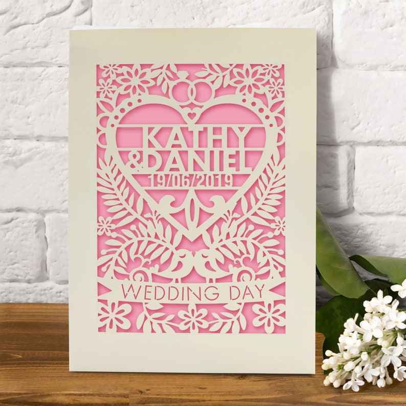 May include: A white card with a pink heart cut out in the centre. The heart has the text "Kathy & Daniel 19/06/2019" inside. The card is decorated with a floral design and the words "Wedding Day" at the bottom.