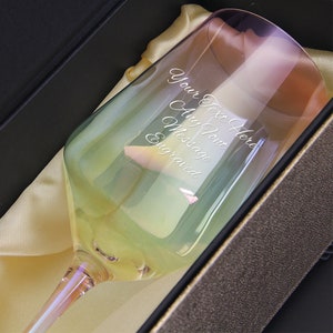 May include: A clear glass wine glass with a rainbow iridescent finish. The glass is in a box and has the text "Your Text Here Any Font Message Engraved" etched on the side.
