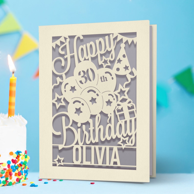 May include: A beige card with a cut-out design of balloons, stars, a gift box, and a party hat. The text "Happy 30th Birthday Olivia" is cut out of the card.