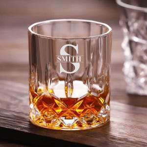 May include: A clear glass whiskey tumbler with a diamond-shaped pattern. The glass is filled with amber liquid and has the letter "S" and the word "SMITH" etched on the front.