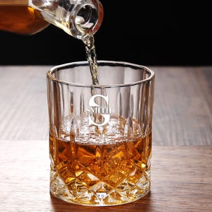 May include: A clear glass whiskey tumbler with a diamond-shaped pattern. The glass is filled with amber liquid and ice. The glass is engraved with the letter "S" and the word "SMITH" underneath.