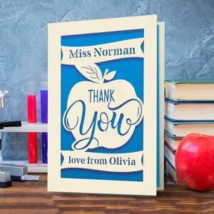 May include: A white card with a blue apple design. The apple has the words "Thank You" inside. The card says "Miss Norman" at the top and "love from Olivia" at the bottom.