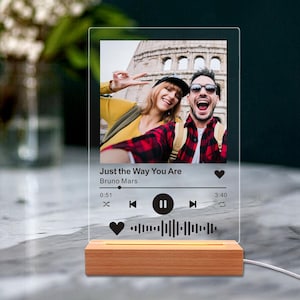 May include: A clear acrylic photo display featuring a selfie of two people, with the song title "Just the Way You Are" by Bruno Mars. The display sits on a wooden base with a warm light.