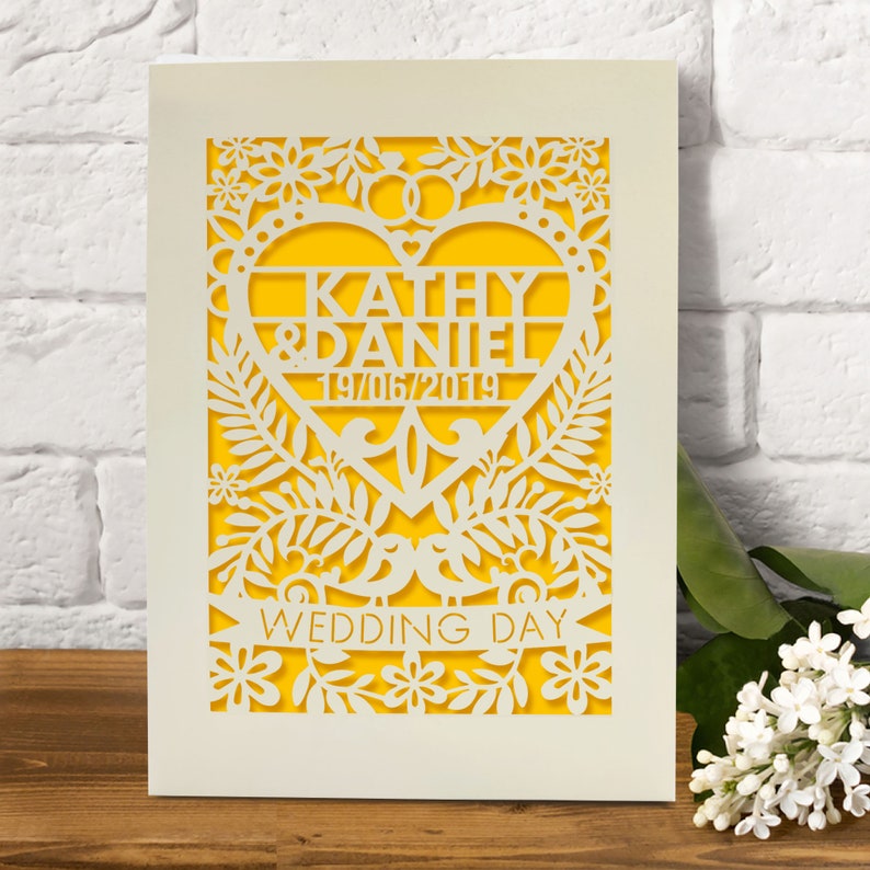 May include: A white card with a yellow background and a heart-shaped cutout. The heart features the names "Kathy & Daniel" and the date "19/06/2019". The card is decorated with a floral design and the words "Wedding Day".