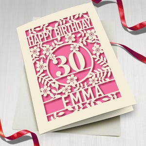 May include: A white card with a pink background and a cut-out design of flowers and leaves. The number 30 is in a circle in the centre of the card. The name Emma is at the bottom of the card. The card says "Happy Birthday".