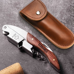 May include: A silver and wood wine opener with a brown leather carrying case. The opener has a wooden handle with the name "Terrance" engraved on it. The opener also has a silver lever with the word "PUSH" and an arrow pointing upwards.