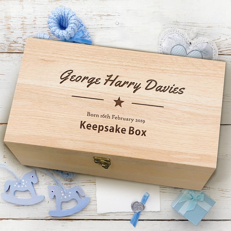 May include: A light wood keepsake box with a personalized inscription: "George Harry Davies Born 16th February 2019 Keepsake Box". The inscription is in a brown font with a star and two lines above and below the text. The box is sitting on a white wooden surface with blue and white decorations.