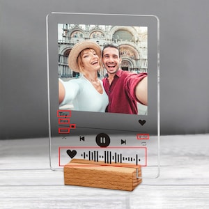 May include: A clear acrylic photo display featuring a couple taking a selfie. The display has a music player interface with a song title, progress bar, and a heart-shaped icon. It rests on a wooden base.