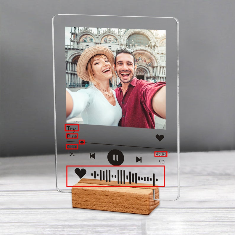 May include: A clear acrylic photo display featuring a couple taking a selfie. The display has a music player interface with a song title, progress bar, and a heart-shaped icon. It rests on a wooden base.