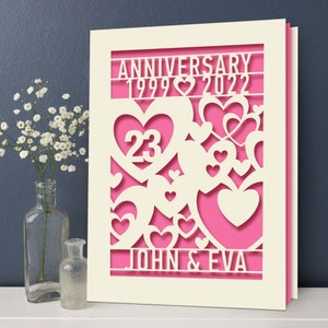 May include: A white card with a pink background and a cut-out design of hearts. The card says "Anniversary 1999 ♥ 2022" and "John & Eva" with the number "23" in a large heart.