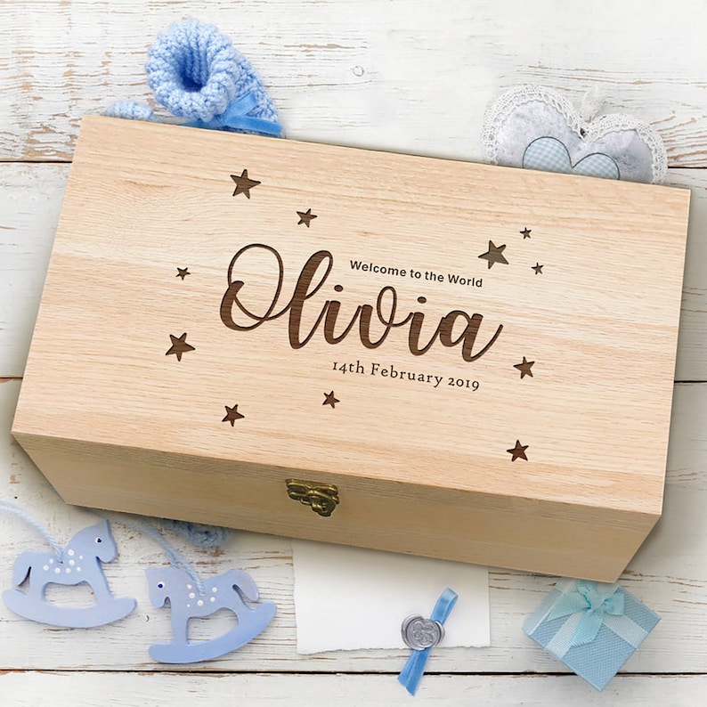May include: A wooden keepsake box with a light wood finish. The lid is engraved with the name "Olivia", the text "Welcome to the World", and the date "14th February 2019". The box is decorated with small stars.