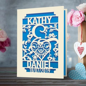 May include: A white card with a blue background and a cutout design of two birds sitting on a branch with a heart in the center. The names "Kathy" and "Daniel" are at the top and bottom of the design, with the date "18/07/2019" below the name "Daniel".