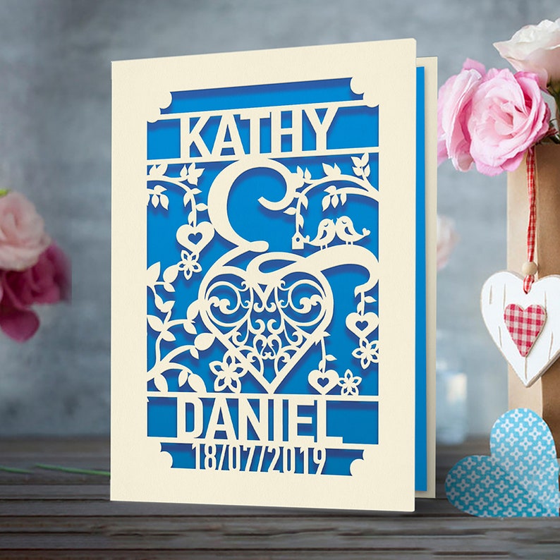 May include: A white card with a blue background and a cutout design of two birds sitting on a branch with a heart in the center. The names "Kathy" and "Daniel" are at the top and bottom of the design, with the date "18/07/2019" below the name "Daniel".