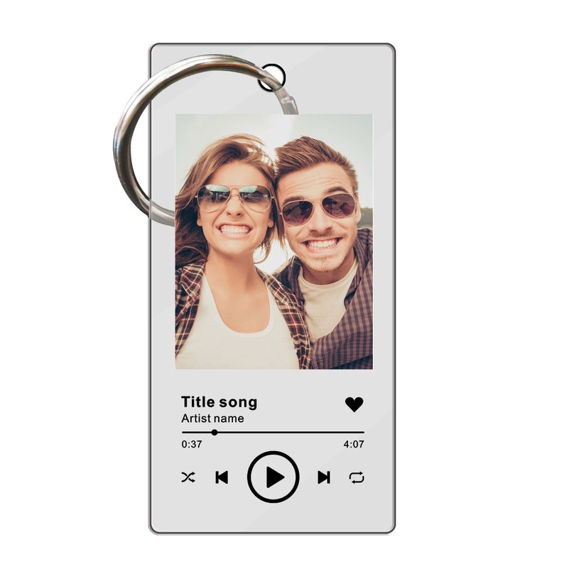 Pu&ograve; includere: Un portachiavi in acrilico bianco con una foto di una coppia con occhiali da sole. Il portachiavi ha un design di lettore musicale con il testo "Title song" e "Artist name". Il lettore musicale ha un pulsante di riproduzione, un'icona a forma di cuore e una barra di avanzamento.