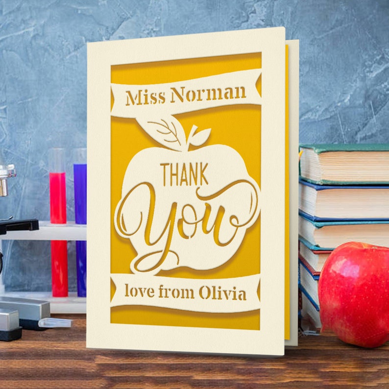 May include: A white card with a yellow apple design. The apple has the words "Thank You" inside. The card says "Miss Norman" and "love from Olivia" on the top and bottom.
