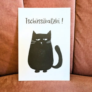 May include: A white card with a black illustration of a grumpy cat and the text "Tsch&uuml;ssikatzki!". The cat has a round body, a curved tail, and white whiskers. The card is displayed on a pink surface.