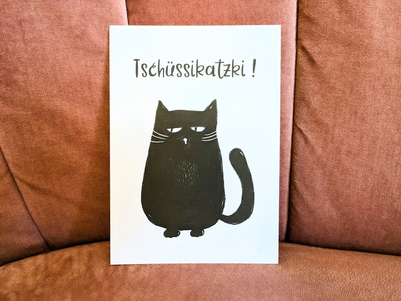 May include: A white card with a black illustration of a grumpy cat and the text "Tsch&uuml;ssikatzki!". The cat has a round body, a curved tail, and white whiskers. The card is displayed on a pink surface.