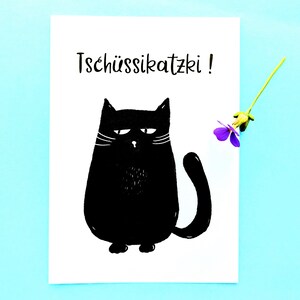 May include: A white card with a black illustration of a grumpy cat and the text "Tsch&uuml;ssikatzki!". A small purple flower with a green stem is placed on the right side of the card against a light blue background.