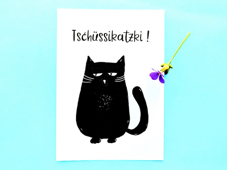 May include: A white card with a black illustration of a grumpy cat and the text "Tsch&uuml;ssikatzki!". A small purple flower with a green stem is placed on the right side of the card against a light blue background.