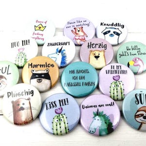 Könnte beinhalten: Eine Sammlung bunter runder Ansteckbuttons mit verschiedenen Illustrationen und Texten. Designs umfassen einen tanzenden Bären, einen Kaktus mit Herz, ein Einhorn und Sätze wie "Kiss Me!" und "Unicorns are real!"