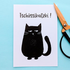 May include: A white card with a black illustration of a grumpy cat and the text "Tsch&uuml;ssikatzki!". A pair of scissors with wooden handles and black blades rests on a light blue surface.