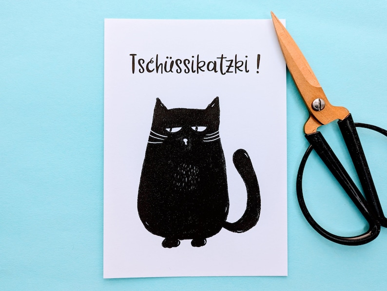 May include: A white card with a black illustration of a grumpy cat and the text "Tsch&uuml;ssikatzki!". A pair of scissors with wooden handles and black blades rests on a light blue surface.