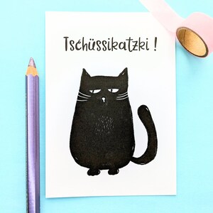May include: A white card with the text "Tsch&uuml;ssikatzki!" and a black cartoon cat illustration. A purple pencil and a roll of pink tape are on a light blue surface.
