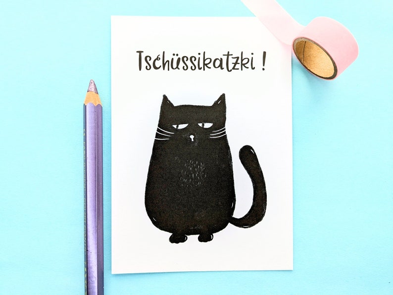 May include: A white card with the text "Tsch&uuml;ssikatzki!" and a black cartoon cat illustration. A purple pencil and a roll of pink tape are on a light blue surface.