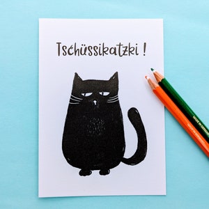 May include: A white card with the German phrase "Tsch&uuml;ssikatzki!" and a black cat illustration. Two colored pencils, orange and green, are on the right side of the card. The background is a light blue surface.
