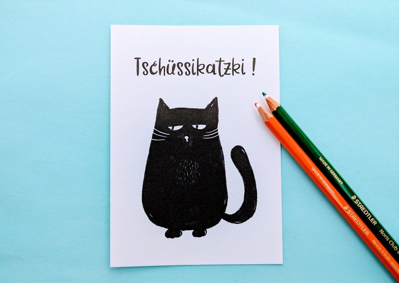 May include: A white card with the German phrase "Tsch&uuml;ssikatzki!" and a black cat illustration. Two colored pencils, orange and green, are on the right side of the card. The background is a light blue surface.