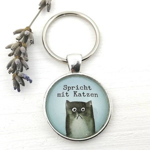 Puede incluir: Un llavero plateado con un colgante redondo que presenta una ilustraci&oacute;n de acuarela de un gato gris con el texto "Spricht mit Katzen" sobre un fondo azul claro. Una ramita de lavanda est&aacute; en el fondo.